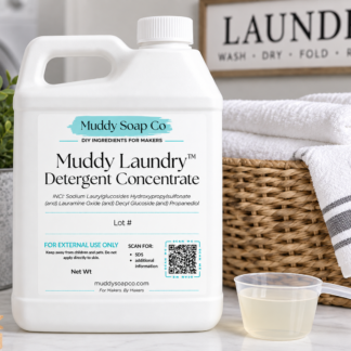 White F-style jug of Muddy Laundry Detergent Concentrate with Muddy Soap Co. label, shown beside a measuring scoop, wicker laundry basket, folded white towels, and clothespins.