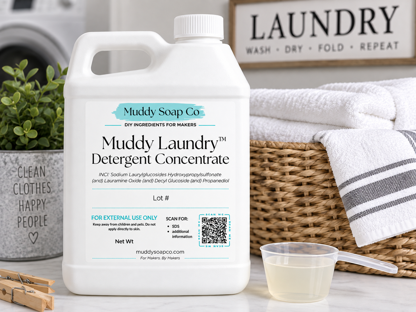White F-style jug of Muddy Laundry Detergent Concentrate with Muddy Soap Co. label, shown beside a measuring scoop, wicker laundry basket, folded white towels, and clothespins.